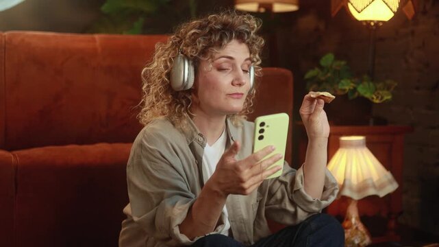 Horizontal video of uninterested curly haired woman snacking on pizza while casually browsing content on smartphone at home. High quality 4k footage