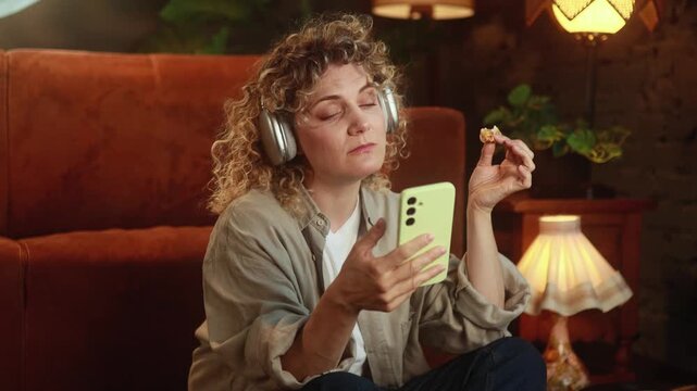 Horizontal video of bored curly haired woman eating pizza and scrolling social media feed on smartphone at home. High quality 4k footage