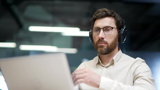 Close-up of focused businessman in wireless headphones working on laptop in modern office. Concentrated professional attending virtual meeting, analyzing data or preparing online business presentation