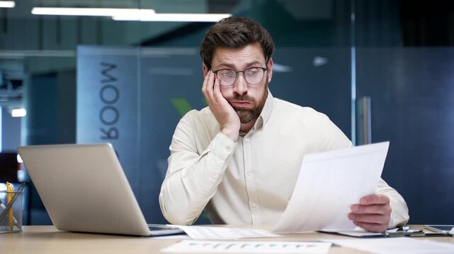 Confused businessman sitting at desk in office struggling with paperwork and looking at laptop screen. Frustrated company owner reviewing documents, facing accounting mistakes or business problems.