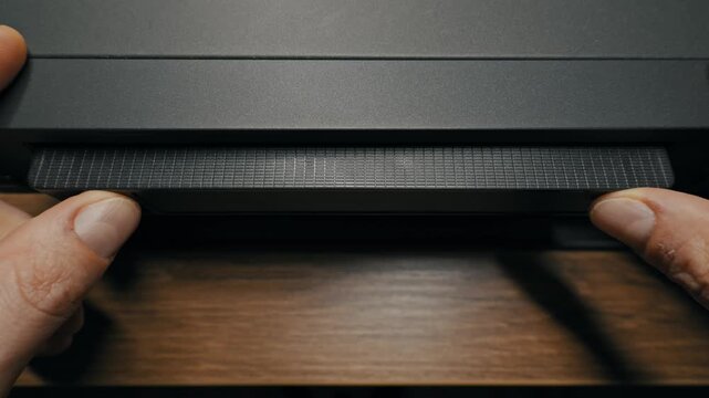Hands insert a black VHS cassette into the slot of a grey VCR player on a wooden surface. Concept of retro technology, 90s nostalgia, and watching old home movies on analog media.