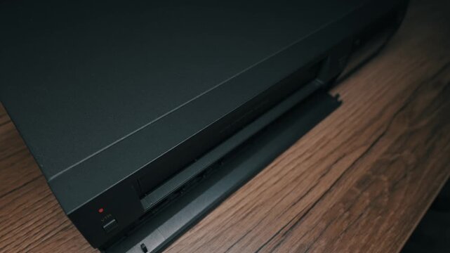 Male hand presses eject button on black VCR player and removes VHS cassette tape. Red indicator light glows against dark matte finish of vintage electronics on wood.