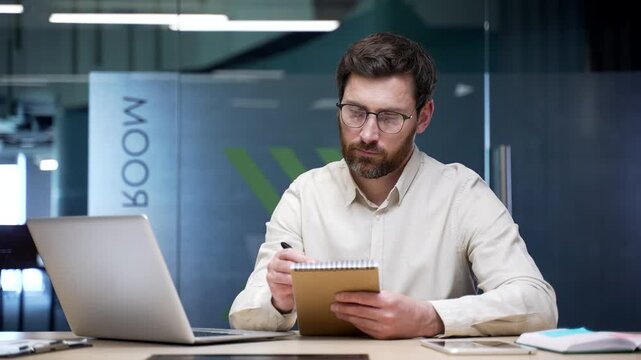 Businessman watching video conference on laptop and taking notes in notebook sitting in office. Focused professional attending virtual training, e-learning course, online seminar for skill development