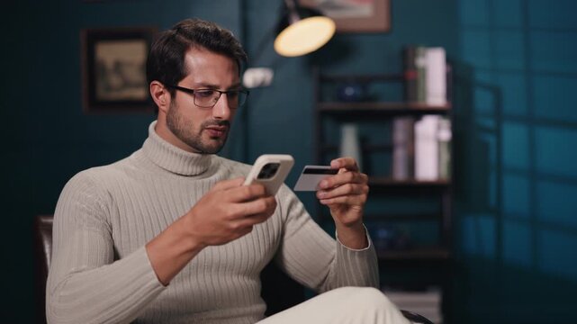Portrait of serious man inserts credit card data into payment app on smartphone in living room. Guy orders goods online on shop website at home. Digital payment