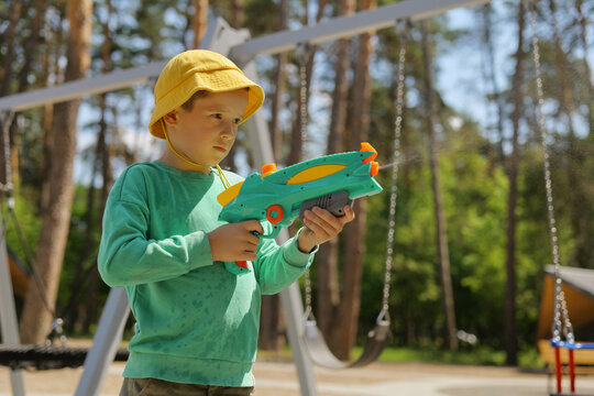 A child stands in a park holding a colorful water toy. The boy wears a yellow hat and green shirt while playing near swings on a sunny day