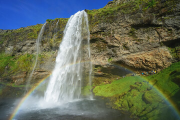A majestic waterfall cascades down a rocky cliff, creating a vibrant rainbow in the mist under a clear blue sky,Seljalandsfoss,Iceland © SHUNJI ZHOU