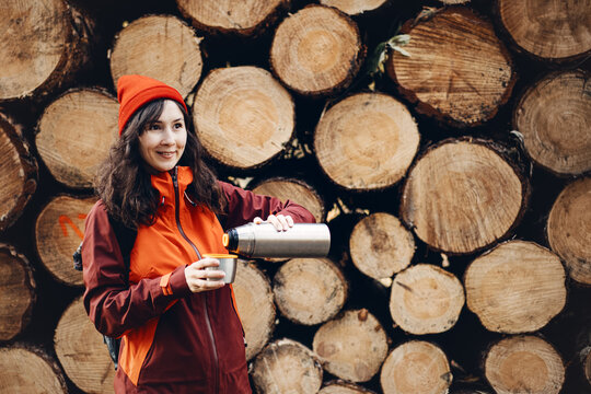 Smiling woman pours drink from metal thermos into cup while standing in front of stacked logs in forest. Hiking break refreshment, outdoor trail pause, recharging energy on trail, Solo travel moments.