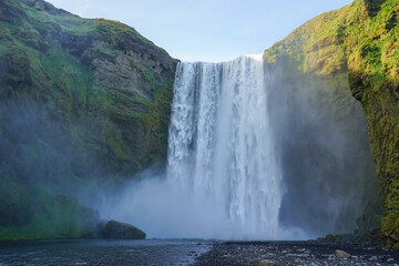 A powerful waterfall cascades down a steep cliff into a misty pool surrounded by lush green mountains,Skogafoss,Iceland © SHUNJI ZHOU