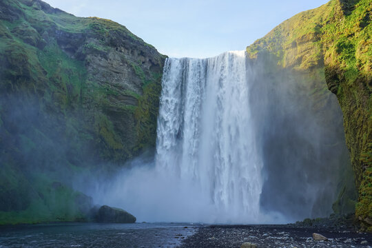 A powerful waterfall cascades down a steep cliff into a misty pool surrounded by lush green mountains,Skogafoss,Iceland