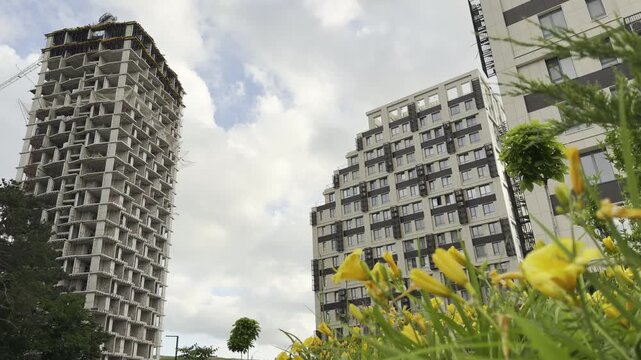 A towering concrete highrise under construction stands near a finished modern apartment building. Bright yellow lilies in foreground create a joyful, refreshing contrast to the growing urban skyline.