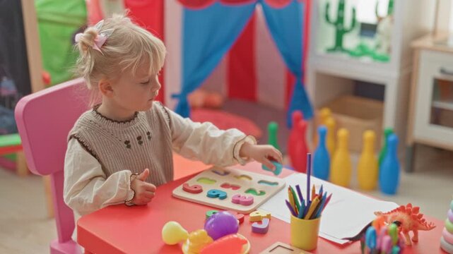 Little blonde girl placing a wooden number puzzle piece with her hand on a table in a colorful preschool; concentration learning.
