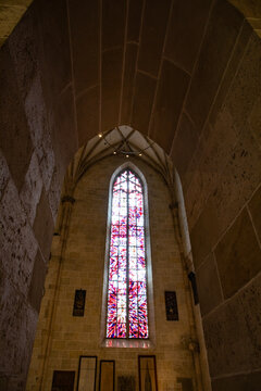 View of a tall gothic stained glass window with vibrant red and purple patterns set in ancient stone walls under a vaulted ceiling in Freiburg im Breisgau, Baden-Wurttemberg, Germany.