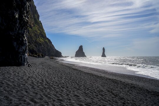 A sandy beach with tall sea stacks and a cliff under a partly cloudy sky,Reynisfjara Black Sand Beach,Iceland