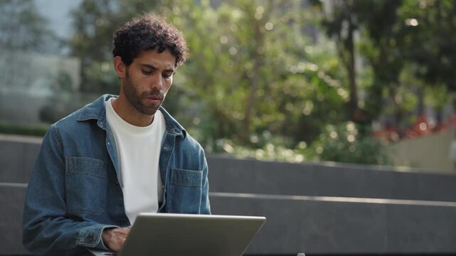 Portrait of focused Hispanic man works on laptop as sits on parapet in park. Bearded Latin guy remote employee writes code on portable computer on city street. Freelancer career