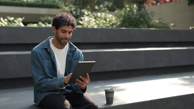 Portrait of cheerful Hispanic man browses internet on digital tablet in urban park. Latin guy in denim jacket scrolls social media via mobile device as sits on stone parapet