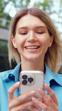 Close-up of satisfied young woman face with long hair and white teeth looking at mobile phone with pleased smile, standing over modern building background