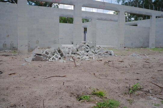 Unfinished cinderblock building at a residential construction site showing concrete block walls, exposed lintels, stacked lumber and gravel forecourt under a blue sky, illustrating building progress a