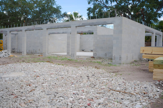 Unfinished cinderblock building at a residential construction site showing concrete block walls, exposed lintels, stacked lumber and gravel forecourt under a blue sky, illustrating building progress a