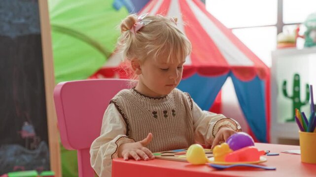 Little blonde girl uses hands to sort colorful plastic toys, craft shapes and pencils on a red table inside a children's playroom building; concentration.