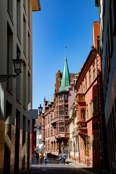 View of the Haus zum Walfisch with its ornate red sandstone facade and a green-spired turret as people walk along the street in Freiburg im Breisgau, Baden-Wurttemberg, Germany.