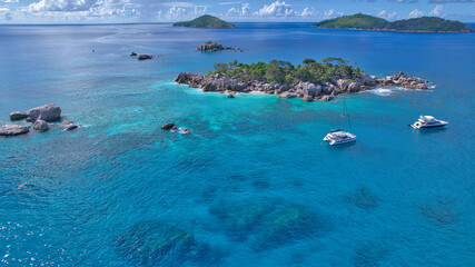Stunning drone shot of Cocos Island, Seychelles, showing a luxury catamaran and boat anchored in clear turquoise water surrounded by granite rocks and coral reefs. © Nils