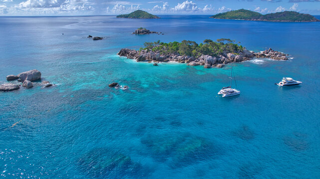 Stunning drone shot of Cocos Island, Seychelles, showing a luxury catamaran and boat anchored in clear turquoise water surrounded by granite rocks and coral reefs.