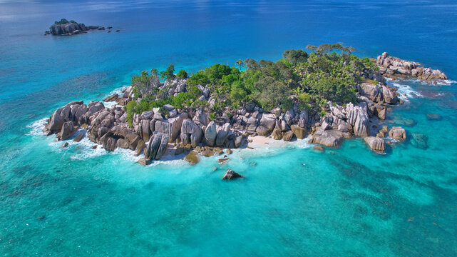Stunning drone shot of Cocos Island, Seychelles, showing a luxury catamaran and boat anchored in clear turquoise water surrounded by granite rocks and coral reefs.