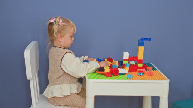 Girl playing with colorful building blocks on a small table, hand reaching to stack bricks in studio; playful curiosity.