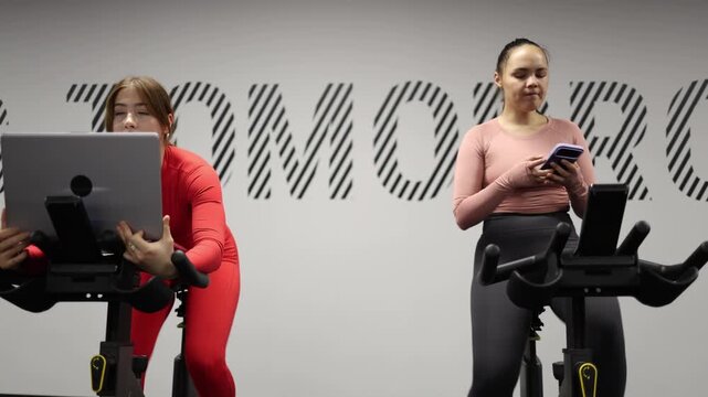 Two women pedal exercise bikes while multitasking one joins an online meeting on a laptop as the other checks messages showing how digital work and constant connectivity blend with fitness
