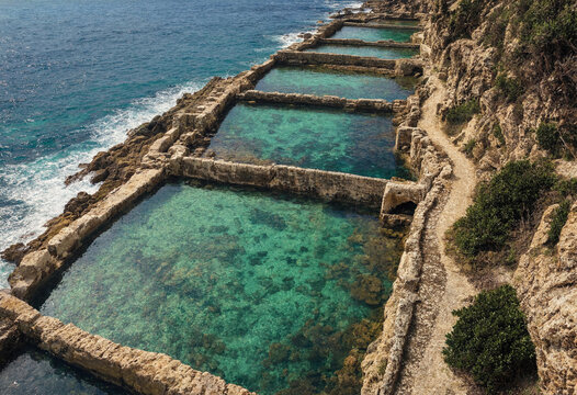 A series of ancient stone salt pans carved into the rocky coastline, filled with clear turquoise seawater under a bright sunny sky by the ocean