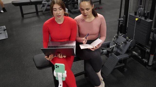 Two women join a work meeting between gym sets showing how hustle culture blends fitness with remote jobs teamwork and nonstop productivity throughout the day