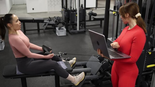 Fitness trainer analyzes workout performance on laptop while client performs seated rowing exercise Combining strength training with data driven coaching