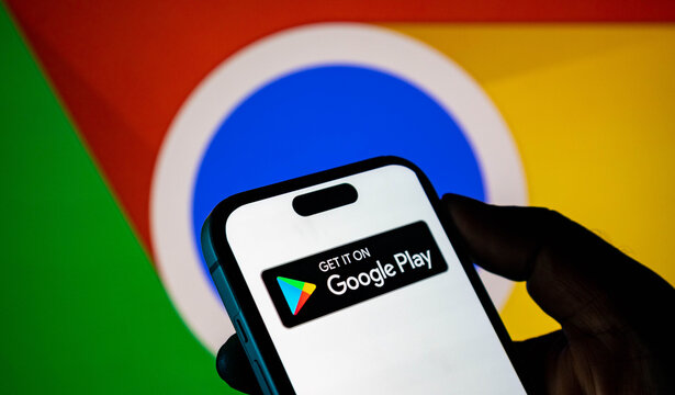 Closeup of a hand holding a smartphone displaying the google play store logo against a google chrome background