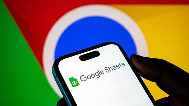 Person holding a smartphone displaying the google sheets app logo with the google chrome logo in the background