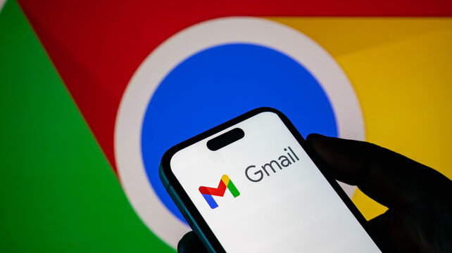 Closeup of a hand holding a smartphone displaying the gmail app logo against a google chrome browser background