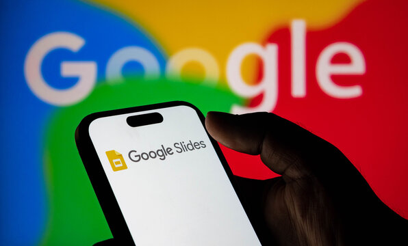 Closeup of a hand holding a smartphone displaying the google slides app logo against a blurred google logo background