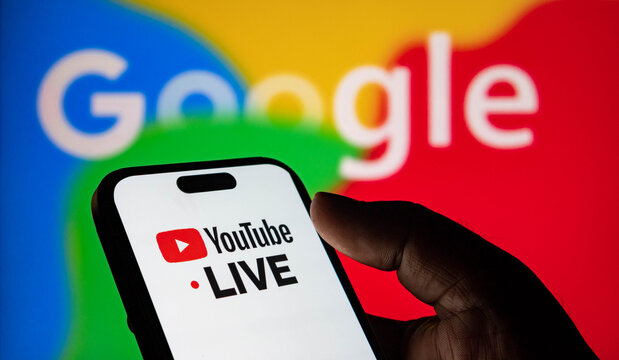 Closeup of a hand holding a smartphone displaying the youtube live logo with the google logo in the background
