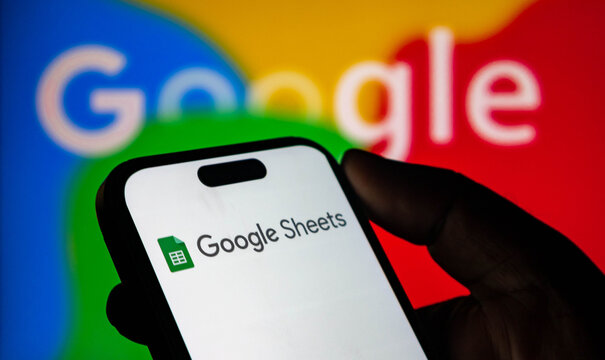Closeup of a hand holding a smartphone displaying the google sheets app logo against a colorful google background