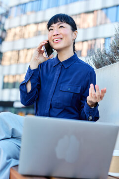 Portrait of a young asian japanese businesswoman woman using a smartphone mobile phone walking down the street, surrounded by moder corporate office buildings