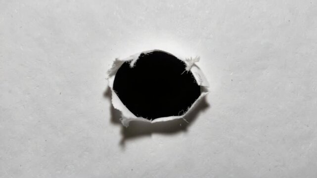 The shows a single bullet hole through paper with visible shrapnel and powder residue surrounding the entry point. The hole is small and darkened due to the impact of the bullet's trajectory.