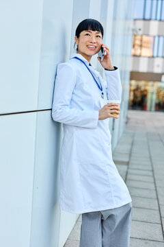 Portrait of an asian female woman doctor or nurse and  health care worker using a smartphone mobile phone holding a cup of coffee   in front of hospital or clinic outside