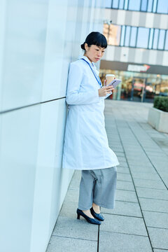 Portrait of an asian female woman doctor or nurse and  health care worker using a smartphone mobile phone holding a cup of coffee   in front of hospital or clinic outside