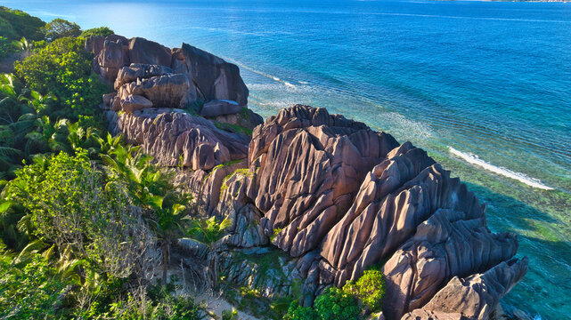 Breathtaking top-down aerial shot of dramatic weathered granite boulders meeting crystal-clear turquoise waters in Anse Grosse Roche, La Digue, Seychelles.