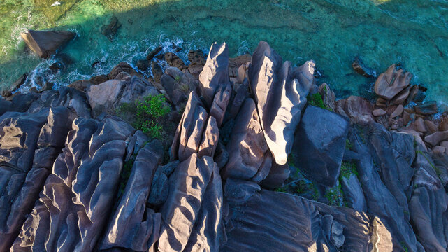 Breathtaking top-down aerial shot of dramatic weathered granite boulders meeting crystal-clear turquoise waters in Anse Grosse Roche, La Digue, Seychelles.