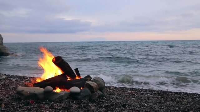 A beach bonfire burns on a rocky shoreline, with large rocks forming a fire pit and logs ablaze, as waves gently lap at the shore in the background.