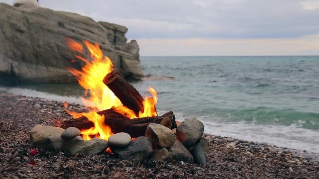 A beach bonfire burns on a rocky shoreline, with large rocks forming a fire pit and logs ablaze, as waves gently lap at the shore in the background.