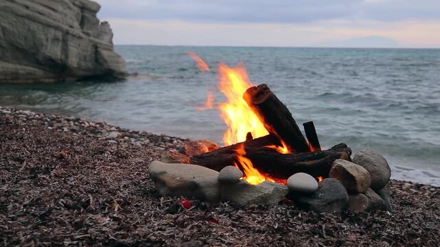 A beach bonfire burns on a rocky shoreline, with large rocks forming a fire pit and logs ablaze, as waves gently lap at the shore in the background.