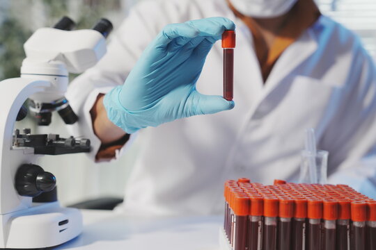 A focused shot of a single blood sample tube held up for visual verification by a scientist, with a microscope and more tubes blurred in the background.