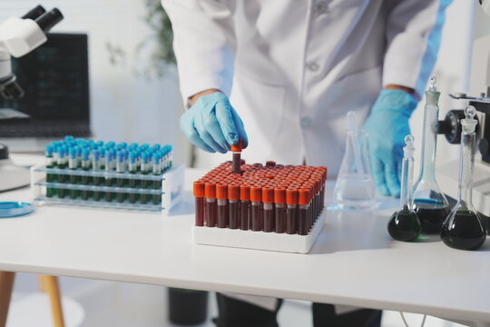 A focused shot of a single blood sample tube held up for visual verification by a scientist, with a microscope and more tubes blurred in the background.