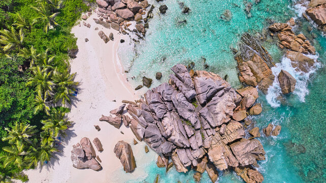 Birds-eye drone shot of giant granite boulders, pristine white sand beach, lush green palm trees, and crystal-clear turquoise waters at Anse Cocos, La Digue, Seychelles. Tropical nature background